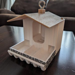May include: A white bird feeder with a brown roof and a metal hook. The feeder has a stone-like design on the bottom and a small opening for birds to enter.