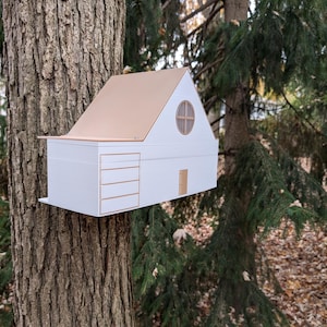 Puede incluir: Un pequeño modelo de casa de madera blanca y marrón con una ventana redonda y una puerta. La casa está unida al tronco de un árbol.