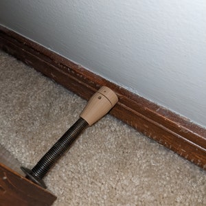 May include: A wooden door stop with a threaded metal rod. The door stop is brown and has a small hole in the top. The door stop is placed against a brown wooden skirting board and a white wall.
