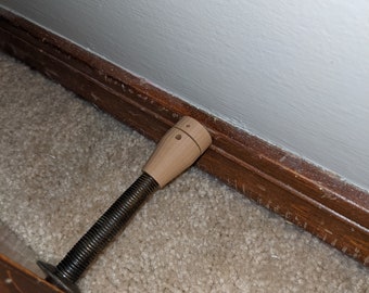 Può includere: Un fermaporta in legno con un'asta di metallo filettata. Il fermaporta è marrone e ha un piccolo foro nella parte superiore. Il fermaporta è posizionato contro uno zoccolo di legno marrone e un muro bianco.