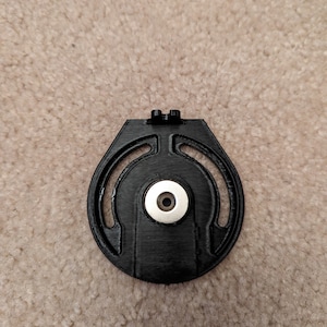 May include: A black plastic 3D printed object with a circular shape and a small silver disc in the center. The object has two small protrusions on the top.