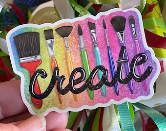 May include: A sticker with a rainbow background and the word "create" in black glitter lettering. There are eight paintbrushes of different colors and sizes arranged across the sticker.