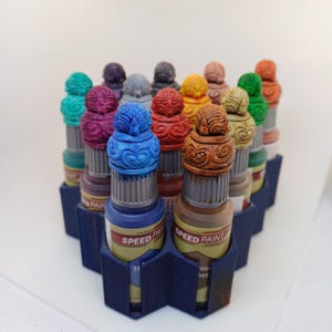 May include: A collection of Speed Paint bottles in various colors, arranged in a blue hexagonal holder. Each bottle has a decorative, ornate cap. The labels read "SPEED PAINT". The colors include blue, purple, green, orange, and brown.