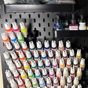 May include: A black pegboard organizer filled with various model paint bottles in a range of colors. Additional supplies include bottles of liquid, plastic containers, and a bag of materials. The paints are organized for easy access.