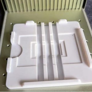 May include: A white plastic test cassette with three test strips inside a light green metal case. The cassette has a rectangular design with a curved cutout and raised edges. The test strips have visible lines.