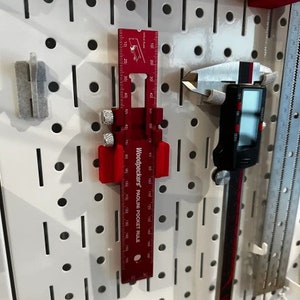 May include: A red Woodpeckers PacLine Pocket Rule with a metal ruler and a sliding measuring tool. The rule is hanging on a white pegboard.