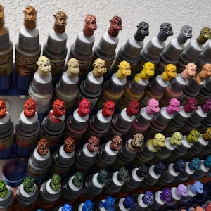 May include: A collection of Speed Paint bottles, each with a unique color and a miniature monster head cap. The bottles are arranged in a blue and white organizer, showcasing a variety of shades, from yellows and reds to greens and purples, for miniature painting.