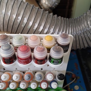 May include: A close-up of a white paint rack with eight small bottles of paint. The bottles are arranged in a row and are labeled with the brand "Citadel" and the color name. The bottles are all different colors, including brown, red, green, and yellow.