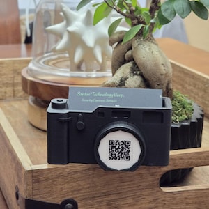 May include: A black camera-shaped business card holder with a white QR code on the lens. The holder is sitting on a wooden tray with a small bonsai tree in a black pot.