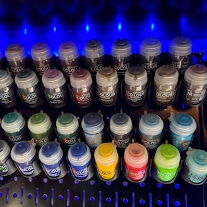 May include: A collection of Citadel Colour paint pots arranged in a black plastic rack. The pots feature various colored lids and labels, including shades of green, blue, yellow, red, and brown, all illuminated under bright blue lighting.