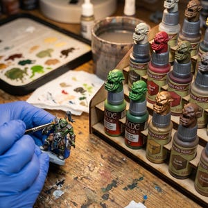 May include: A person wearing blue gloves paints a miniature figurine with a brush. The figurine is a green and gray orc. Paint bottles with orc head toppers and paint colors are on a wooden table, along with a paint palette and a water container.