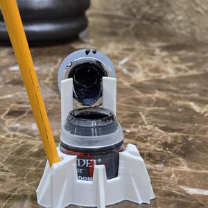 May include: A black paint bottle is held in a white 3D-printed holder. The bottle is open, revealing the dark paint inside. A yellow pencil is leaning against the bottle. The bottle has the text "DEL SE DON".