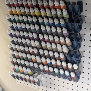 May include: A pegboard with a variety of small bottles of paint organized in rows. The bottles are arranged in a rainbow pattern, with the colors transitioning from red to violet. The bottles are held in place by blue plastic holders.