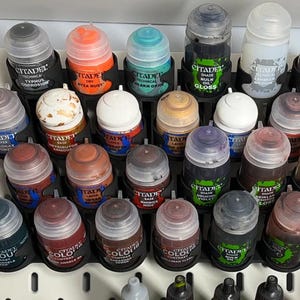May include: A collection of Citadel paint pots in various colors, including orange, blue, and brown, arranged in a black organizer. The pots are labeled with product names and are designed for miniature painting.