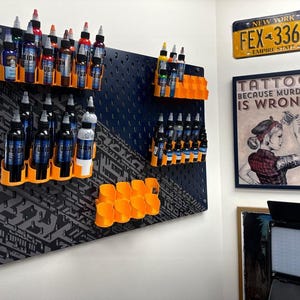 May include: A black pegboard with orange and black ink bottle holders. The pegboard is covered in a black and white pattern. There are several rows of ink bottles in the holders. The pegboard is mounted on a white wall. There is a sign that says "Tattoo because murder is wrong" on the wall.