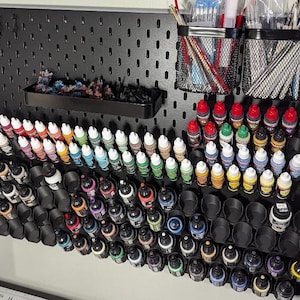 May include: A black pegboard organizer with various art supplies. It features rows of paint bottles in multiple colors, black mesh containers holding paint brushes, and a small tray with miniature figurines. The setup is designed for art and hobby enthusiasts.