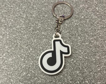 May include: A white and black keychain featuring the TikTok logo. The charm is in the shape of a musical note with a black interior and a white outline. The keychain has a silver metal ring and chain.