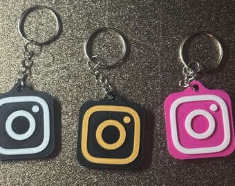 May include: Three keychains with different colored Instagram logos. The logos are black with a white outline, yellow with a black outline, and pink with a white outline.