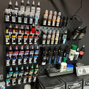 May include: A black pegboard wall with rows of tattoo ink bottles in various colors, including black, white, red, blue, green, yellow, and orange. The bottles are organized by color and brand. There are also some cleaning supplies and other tattoo equipment on the wall.