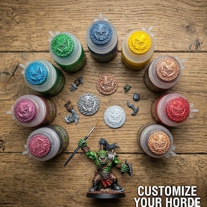 May include: A collection of eight paint pots with embossed orc face lids in various colors, four circular orc face tokens, small miniature parts, a paintbrush, and a painted green orc miniature. Text reads: CUSTOMIZE YOUR HORDE.
