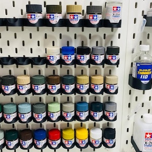 May include: A white pegboard wall with rows of small paint bottles in various colors, organized in black plastic holders. There are also bottles of paint thinner and a few paintbrushes.