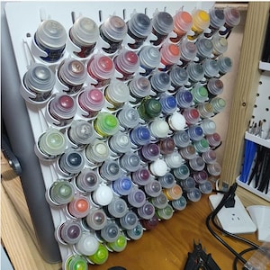 May include: A white paint bottle organizer filled with numerous small paint bottles. The bottles are arranged in a grid, showcasing a variety of colors, including red, green, blue, and yellow. The organizer is mounted on a wall, providing a neat storage solution.