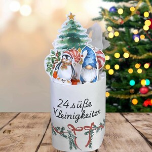 Könnte beinhalten: Ein weißer Weihnachtskalender mit einem Pinguin-, Wichtel- und Weihnachtsbaum-Design. Der Kalender trägt den Text "24 süße Kleinigkeiten" und ist mit roten Schleifen und grünen Girlanden verziert. Im Hintergrund ist ein Weihnachtsbaum zu sehen.