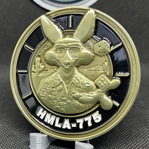 Puede incluir: Una moneda de desafío de color dorado con fondo negro. La moneda presenta un zorro de dibujos animados con gafas de sol y una camisa hawaiana. El zorro sostiene un par de dados. El texto "HMLA-775" está impreso en la moneda.