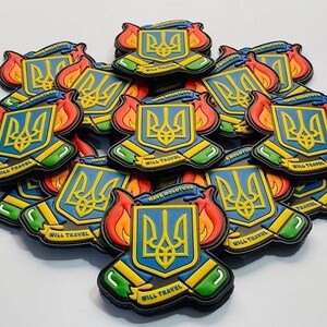 May include: A collection of colorful, layered pins featuring the Ukrainian coat of arms, a trident, in blue and yellow. The pins have a flame design in red, orange, and yellow. Text on the pins reads "WILL TRAVEL" and "HAVE MOLOTOVS".