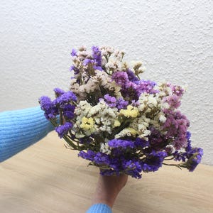 Könnte beinhalten: Ein Strauß getrockneter Blumen in Lila, Weiß und Gelb. Die Blumen werden in einer Hand gehalten, die einen hellblauen Pullover trägt. Der Hintergrund ist eine weiße, strukturierte Wand und eine hellbraune Holzoberfläche.