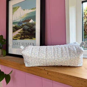May include: A rectangular pencil case with a light blue floral pattern, featuring a zip closure. It rests on a wooden windowsill. A framed poster of a coastal scene hangs on the pink wall behind it.
