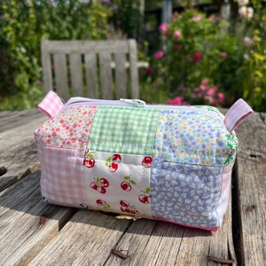 K&ouml;nnte beinhalten: Eine Rei&szlig;verschlusstasche aus Patchwork-Stoff mit einem floralen Muster in Rosa, Gr&uuml;n, Wei&szlig; und Blau. Die Tasche liegt auf einer Holzoberfl&auml;che.