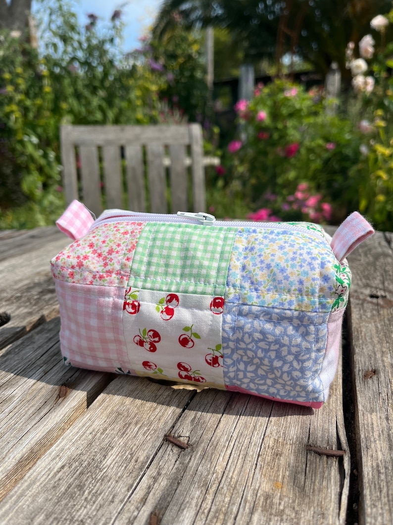 K&ouml;nnte beinhalten: Eine Rei&szlig;verschlusstasche aus Patchwork-Stoff mit einem floralen Muster in Rosa, Gr&uuml;n, Wei&szlig; und Blau. Die Tasche liegt auf einer Holzoberfl&auml;che.