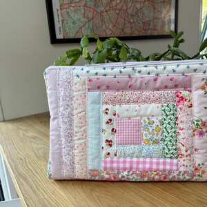 May include: A pink and white patchwork fabric zippered pouch with a log cabin quilt pattern. The pouch is made of various floral and geometric prints.