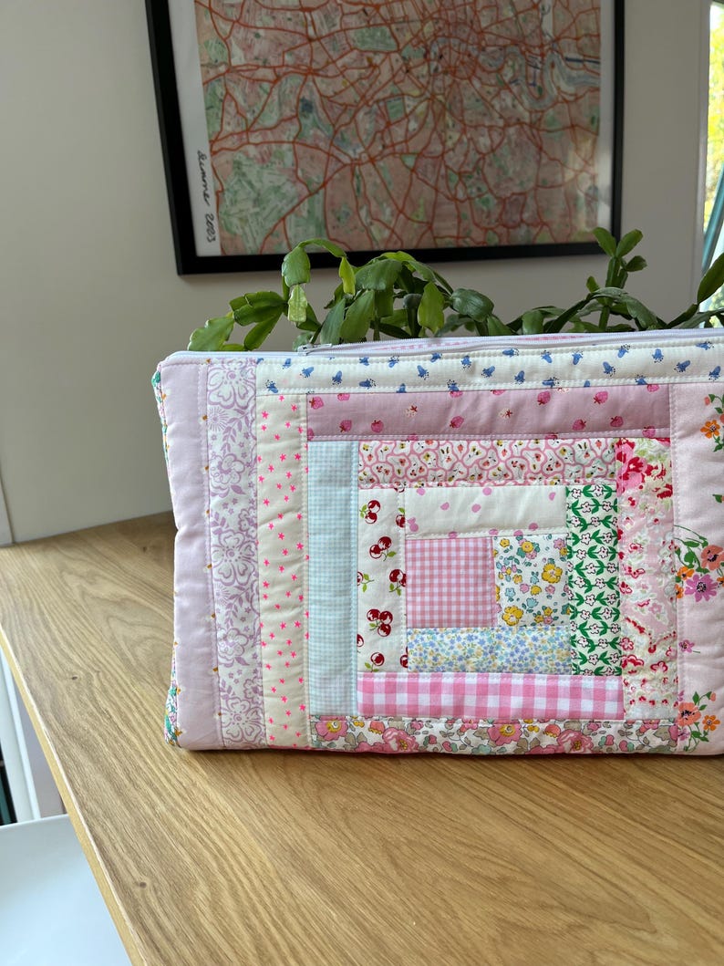 May include: A pink and white patchwork fabric zippered pouch with a log cabin quilt pattern. The pouch is made of various floral and geometric prints.