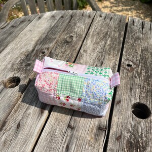 K&ouml;nnte beinhalten: Eine kleine, gepatchte Stofftasche mit Rei&szlig;verschluss und einem rosa, gr&uuml;nen und blauen Blumenmuster. Die Tasche liegt auf einem Holztisch.