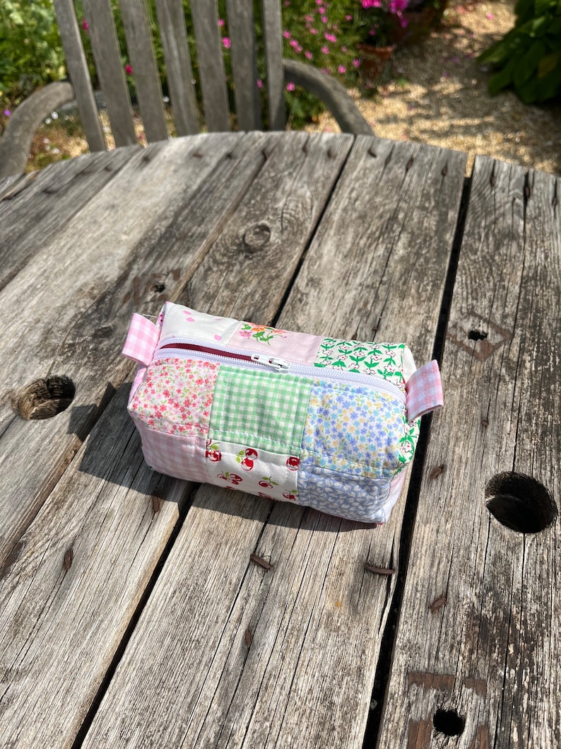 K&ouml;nnte beinhalten: Eine kleine, gepatchte Stofftasche mit Rei&szlig;verschluss und einem rosa, gr&uuml;nen und blauen Blumenmuster. Die Tasche liegt auf einem Holztisch.