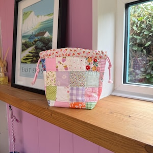 May include: A small drawstring pouch featuring a patchwork design made of various fabric squares in shades of pink, green, purple, and white with floral, gingham, and dotted patterns. The top edge has a cream fabric with a red floral print.