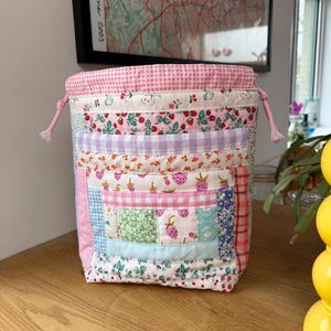 May include: A fabric drawstring pouch featuring a patchwork design of various pastel-coloured cotton prints, including strawberries, gingham checks, and floral patterns. The bag has a pink gingham top border and matching pink drawstring cords.