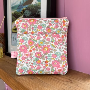 Könnte beinhalten: Eine floral bedruckte Tasche mit einem rosa Reißverschluss. Die Tasche hat einen weißen Hintergrund mit rosa, blauen und gelben Blumen. Ein kleines rosa-weißes Vichy-Band ist an der Seite befestigt. Die Tasche liegt auf einer Holzoberfläche.
