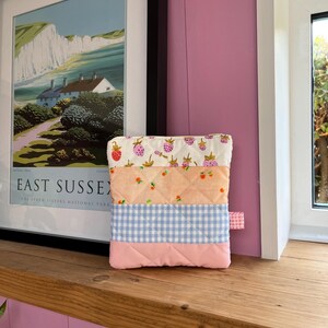 Pu&ograve; includere: Un astuccio in tessuto trapuntato con un design patchwork. La sezione superiore presenta uno sfondo bianco con fragole rosse, seguito da tessuto color pesca con fragole, un motivo a quadretti blu e una base rosa. &Egrave; attaccata una piccola linguetta rosa.