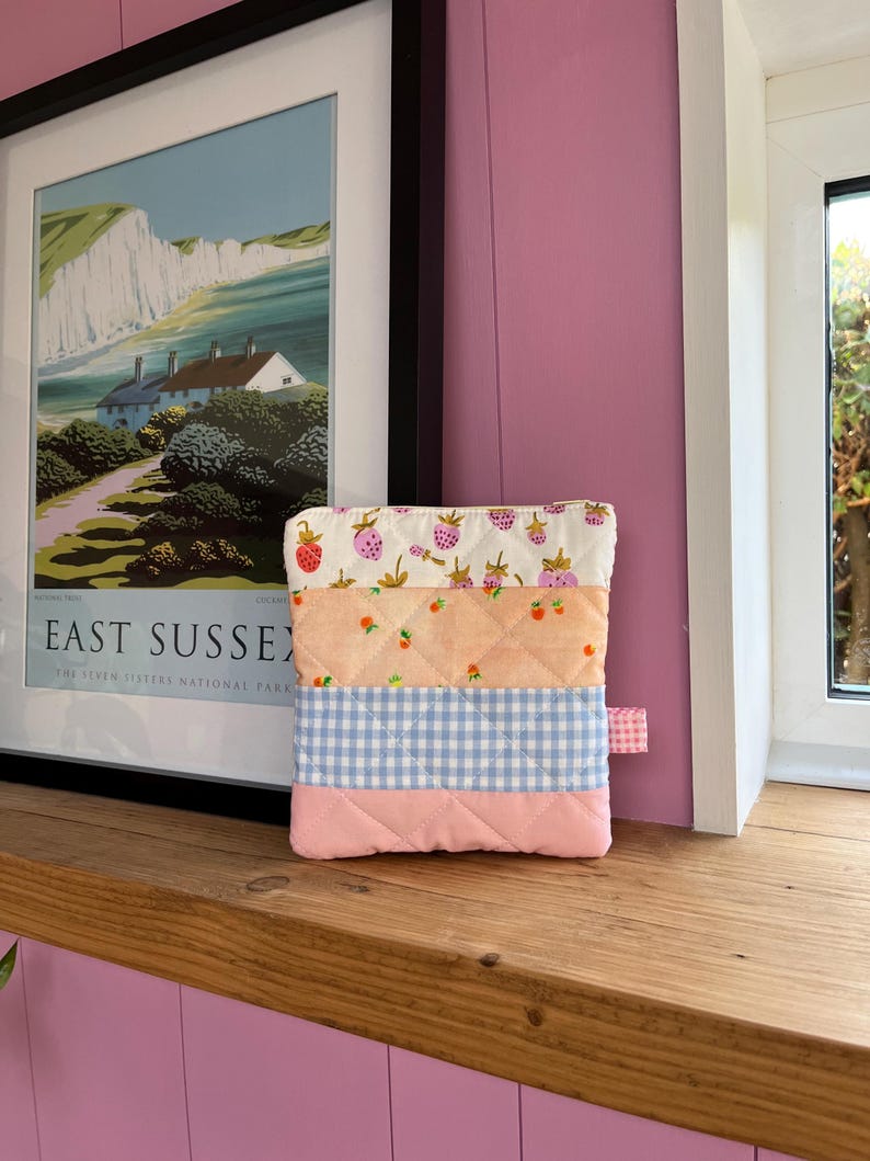 Pu&ograve; includere: Un astuccio in tessuto trapuntato con un design patchwork. La sezione superiore presenta uno sfondo bianco con fragole rosse, seguito da tessuto color pesca con fragole, un motivo a quadretti blu e una base rosa. &Egrave; attaccata una piccola linguetta rosa.