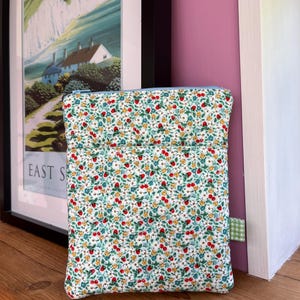 May include: A rectangular fabric pouch featuring a repeating pattern of small red strawberries, yellow flowers, and green leaves on a white background. The pouch has a light blue zipper closure at the top and a small green and white chequered fabric tab.