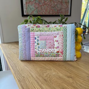 May include: A quilted laptop sleeve with a patchwork design featuring pink, purple, and green floral fabrics. The sleeve has a zipper closure and is made of cotton.