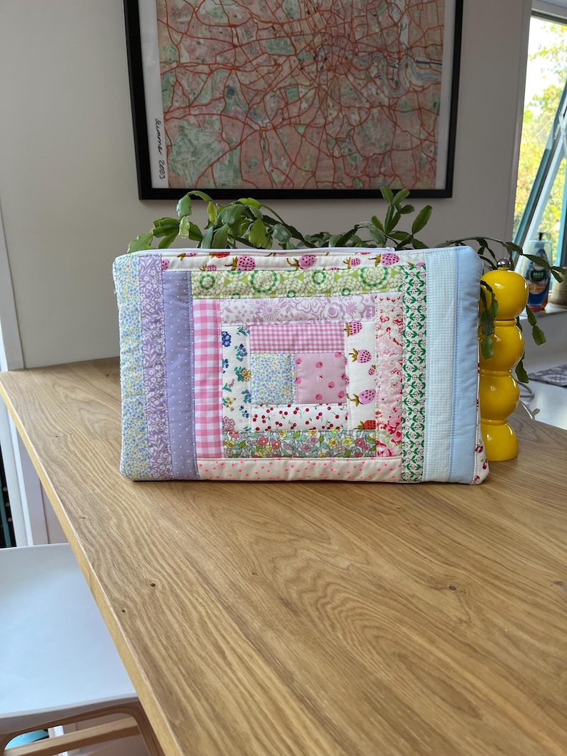 May include: A quilted laptop sleeve with a patchwork design featuring pink, purple, and green floral fabrics. The sleeve has a zipper closure and is made of cotton.