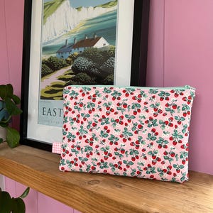 May include: A rectangular pink fabric pouch with a strawberry pattern, featuring red strawberries, green leaves, and white flowers. The pouch has a light green zipper and a small gingham tag. A framed landscape print is in the background.