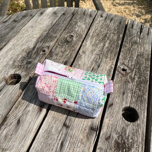 K&ouml;nnte beinhalten: Eine kleine, gepatchte Stofftasche mit Rei&szlig;verschluss in einem rosa, gr&uuml;nen, wei&szlig;en und blauen Blumenmuster. Die Tasche liegt auf einem verwitterten Holztisch.