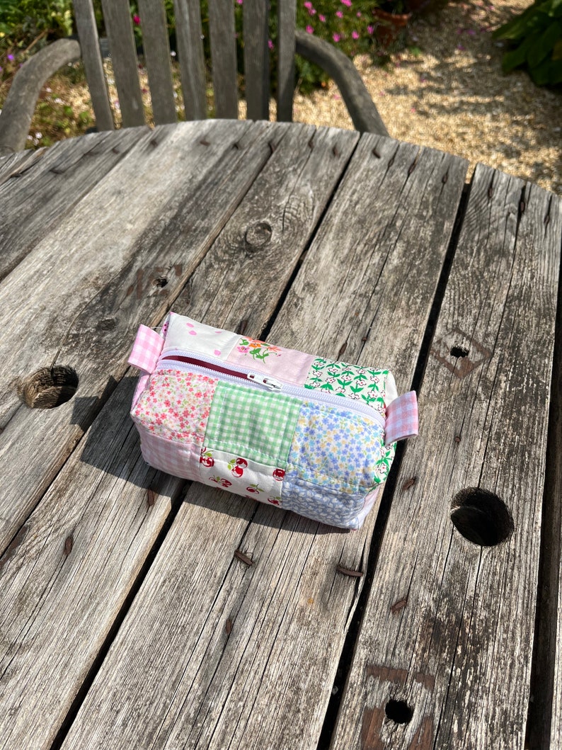 K&ouml;nnte beinhalten: Eine kleine, gepatchte Stofftasche mit Rei&szlig;verschluss in einem rosa, gr&uuml;nen, wei&szlig;en und blauen Blumenmuster. Die Tasche liegt auf einem verwitterten Holztisch.