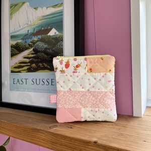 Pu&ograve; includere: Un piccolo astuccio trapuntato con cerniera gialla e tirante a quadretti rosa. Il design patchwork presenta fragole, pois e motivi floreali nei toni del rosa e del bianco. L'astuccio &egrave; esposto su una mensola di legno.