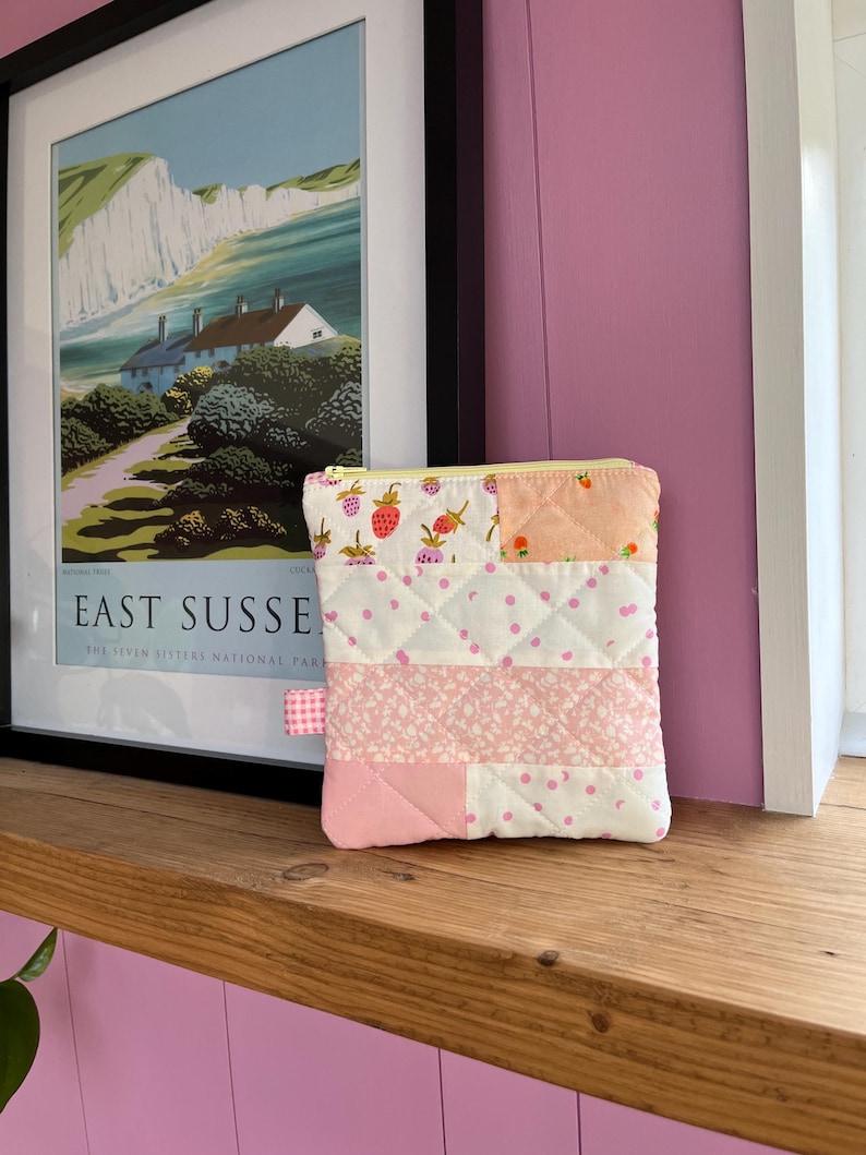 Pu&ograve; includere: Un piccolo astuccio trapuntato con cerniera gialla e tirante a quadretti rosa. Il design patchwork presenta fragole, pois e motivi floreali nei toni del rosa e del bianco. L'astuccio &egrave; esposto su una mensola di legno.